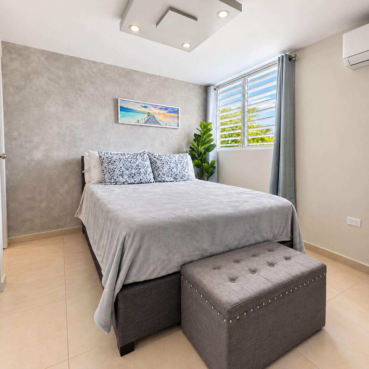 Un dormitorio con una cama, un banco tapizado gris, una ventana con cortinas, una planta en maceta y un cuadro en la pared.