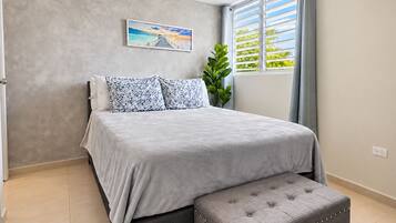 Un dormitorio con una cama, un banco tapizado gris, una ventana con cortinas, una planta en maceta y un cuadro en la pared.