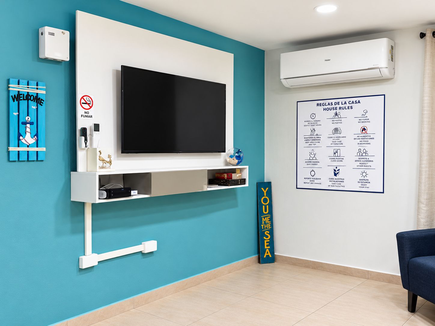 Ein Hotelzimmer mit einem großen Flachbildfernseher an einer weißen Wand, einer Wandklimaanlage, einem 'Welcome'-Schild, einem 'Rauchen verboten'-Schild und einem Aushang mit den Hausregeln.