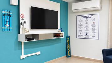 Ein Hotelzimmer mit einem großen Flachbildfernseher an einer weißen Wand, einer Wandklimaanlage, einem 'Welcome'-Schild, einem 'Rauchen verboten'-Schild und einem Aushang mit den Hausregeln.