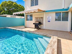 A swimming pool with clear blue water, a barbecue area, and a pool rules sign.