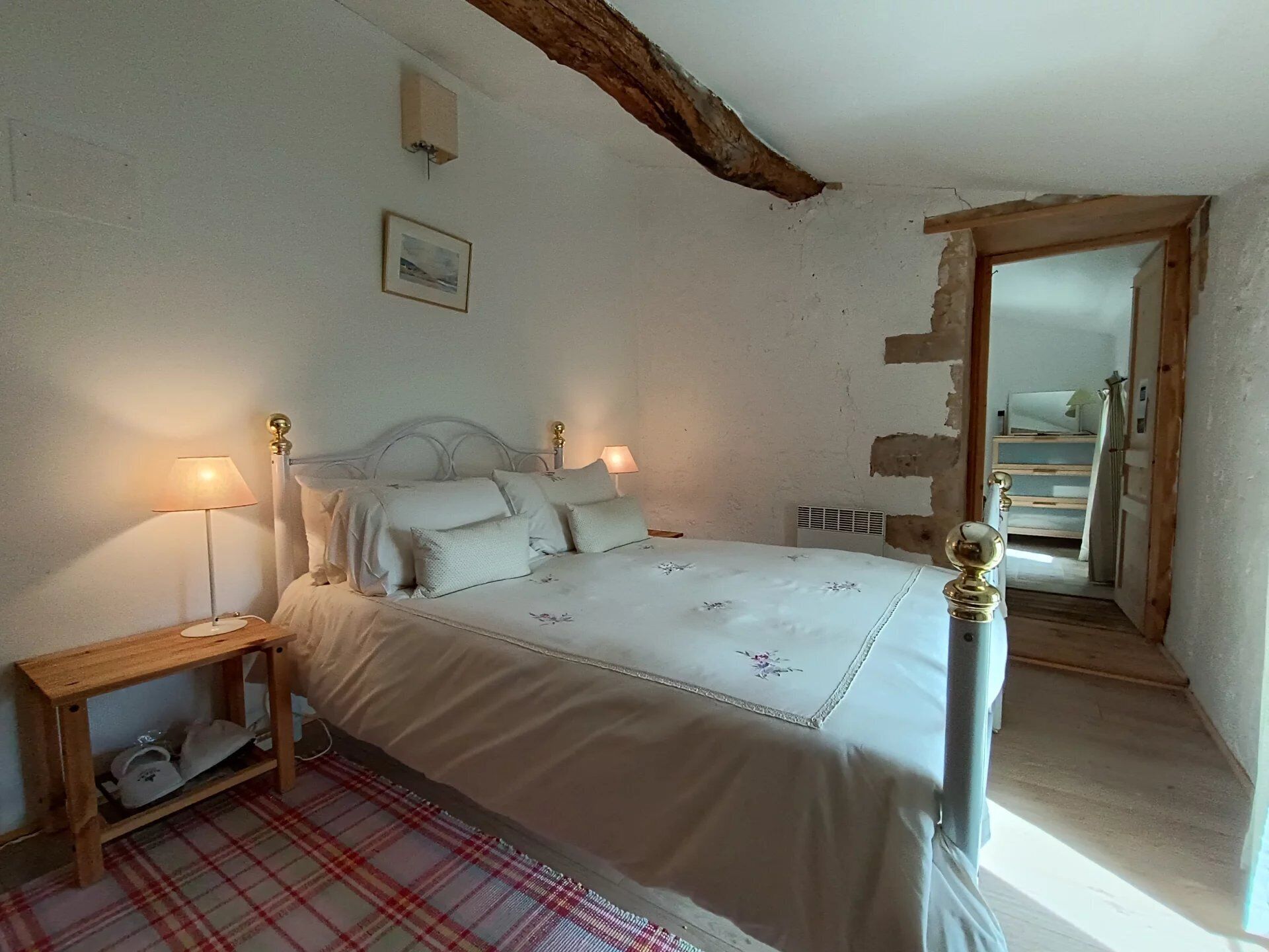 A bedroom with a bed, bedside lamps, a wooden nightstand, a red plaid rug, a framed picture on the wall, and a mirror in the corner.