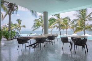Ein großzügiger Außenbereich mit Holztisch und -stühlen, umgeben von Palmen und mit Blick auf einen Pool und den Ozean.