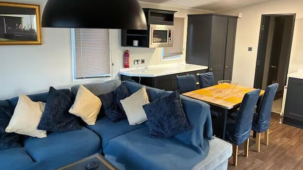 Una sala de estar moderna con un sofá modular, una mesa de comedor con sillas, una zona de cocina y una lámpara colgante negra de gran tamaño.