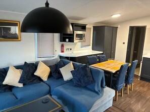 A modern living room with a sectional sofa, a dining table with chairs, a kitchen area, and a large black pendant light. - Cabin Sleeps 6 With Pets (New Romney)