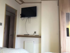 A hotel room with a bed, a TV mounted on the wall, a radiator, and a wooden door. - Cabin Sleeps 6 With Pets (New Romney)