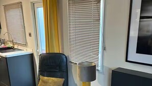 Una sala de estar moderna con un sillón de cuero negro, una mesita auxiliar con una lámpara, una chimenea y una zona de cocina con un lavamanos y persianas.