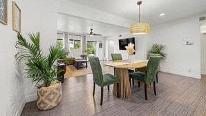 Un vaste espace dédié à la salle à manger, avec une table en bois et des chaises recouvertes d’un tissu vert, le tout agrémenté de plantes en pot et d’un grand luminaire suspendu en forme de suspension tissée.