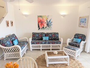 Ein Zimmer mit einer weißen Rattan-Sofagarnitur, einem Glastisch und einem gewebten Teppich.
