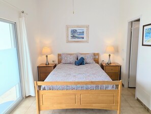 Ein Schlafzimmer mit einem hölzernen Bett, zwei Nachttischen, einem Fenster mit Vorhängen, einer Tür und einem gerahmten Bild an der Wand.
