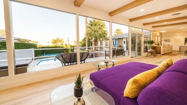 Ein modernes Hotelzimmer mit großen Fenstern, Blick auf den Pool, einem violetten Sofa und einem kleinen Tisch mit Pflanze.
