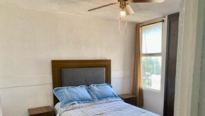 Un dormitorio con cama, dos mesitas de noche, un ventilador de techo y una ventana.
