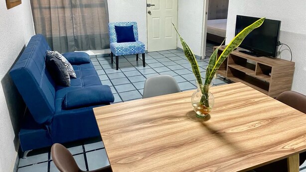 Una habitación con un sofá azul, una mesa de comedor de madera con un florero y plantas, un televisor y una pequeña silla.