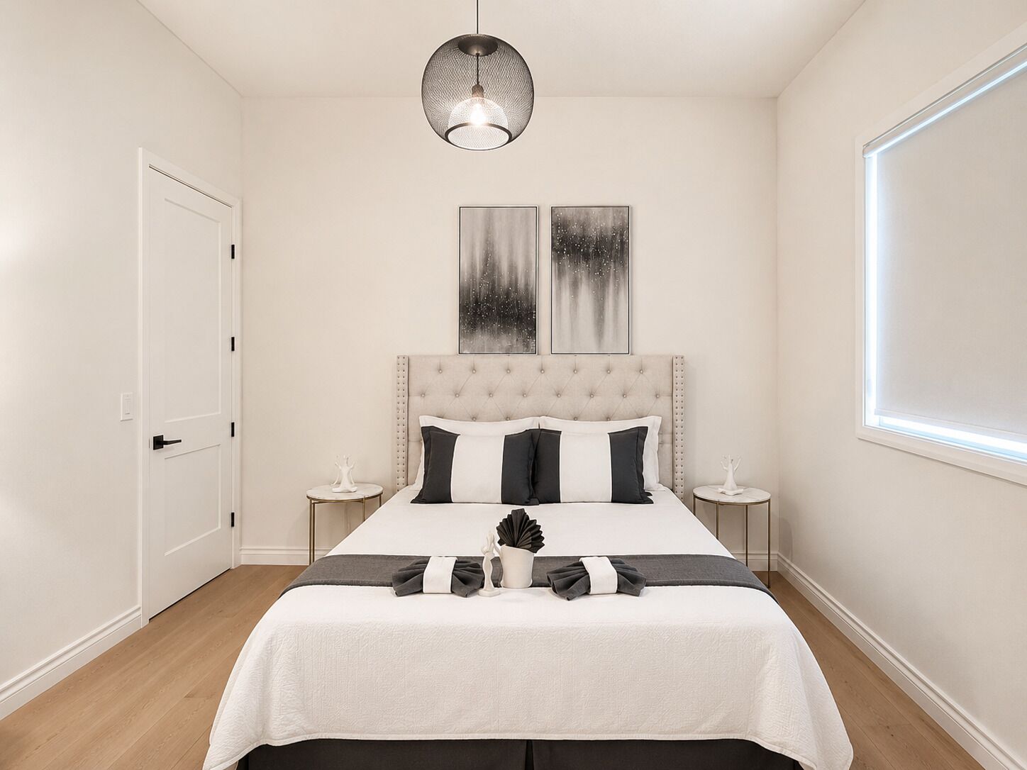 Un dormitorio con cabecera capitonada, cama con colcha blanco y negro, dos mesitas de noche con adornos blancos y un ventanal con persianas.