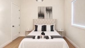 Un dormitorio con cabecera capitonada, cama con colcha blanco y negro, dos mesitas de noche con adornos blancos y un ventanal con persianas.