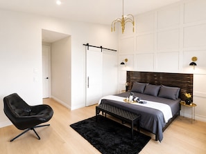 A modern bedroom with a bed, a black chair, a bench, a black rug, and a wall-mounted light fixture.
