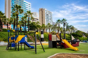 Ein Spielplatz mit Rutschen und Klettergerüsten, umgeben von Palmen und Gebäuden.