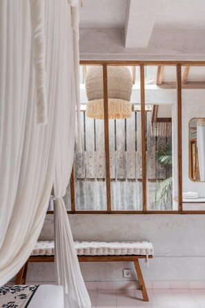 Ein Schlafzimmer mit einem Bett, einer Bank, einer Pendelleuchte, einer Holzwand und einem Fenster mit Blick auf einen Zaun und Pflanzen.