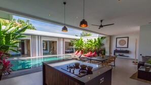 A modern kitchen with a central island, a dining area with chairs, a ceiling fan, and a view of a swimming pool. - Cozy 3BR Villa w  Private Pool near Berawa Beach (Kecamatan Kuta Utara)