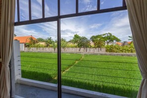 A window with a view of a green field, possibly a rice field, with a clear sky and some trees in the background. - Loft Style Villa with Pool and Ricefield View (Seminyak)