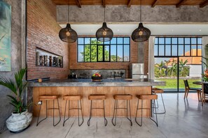A modern kitchen with a large island, bar stools, hanging lights, and a view of greenery outside. - Loft Style Villa with Pool and Ricefield View (Seminyak)
