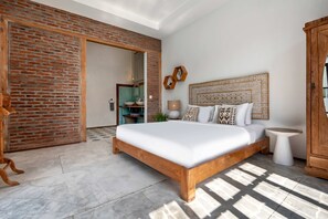 A bedroom with a wooden bed, a brick wall, and a large window casting shadows on the floor. - Loft Style Villa with Pool and Ricefield View (Seminyak)