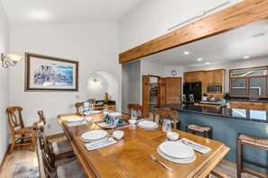 Un coin repas avec une table en bois dressée pour quatre personnes, une cuisine avec des meubles en bois et, au mur, un tableau représentant un paysage urbain encadré.