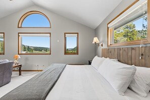 Une chambre à coucher avec un grand lit, deux fenêtres donnant sur un paysage, une table de chevet avec une lampe et une tête de lit en bois.