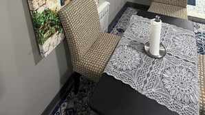Une pièce avec un mur au style rustique, une fenêtre avec des volets bleus, deux chaises à motifs, une nappe en dentelle et un radiateur.