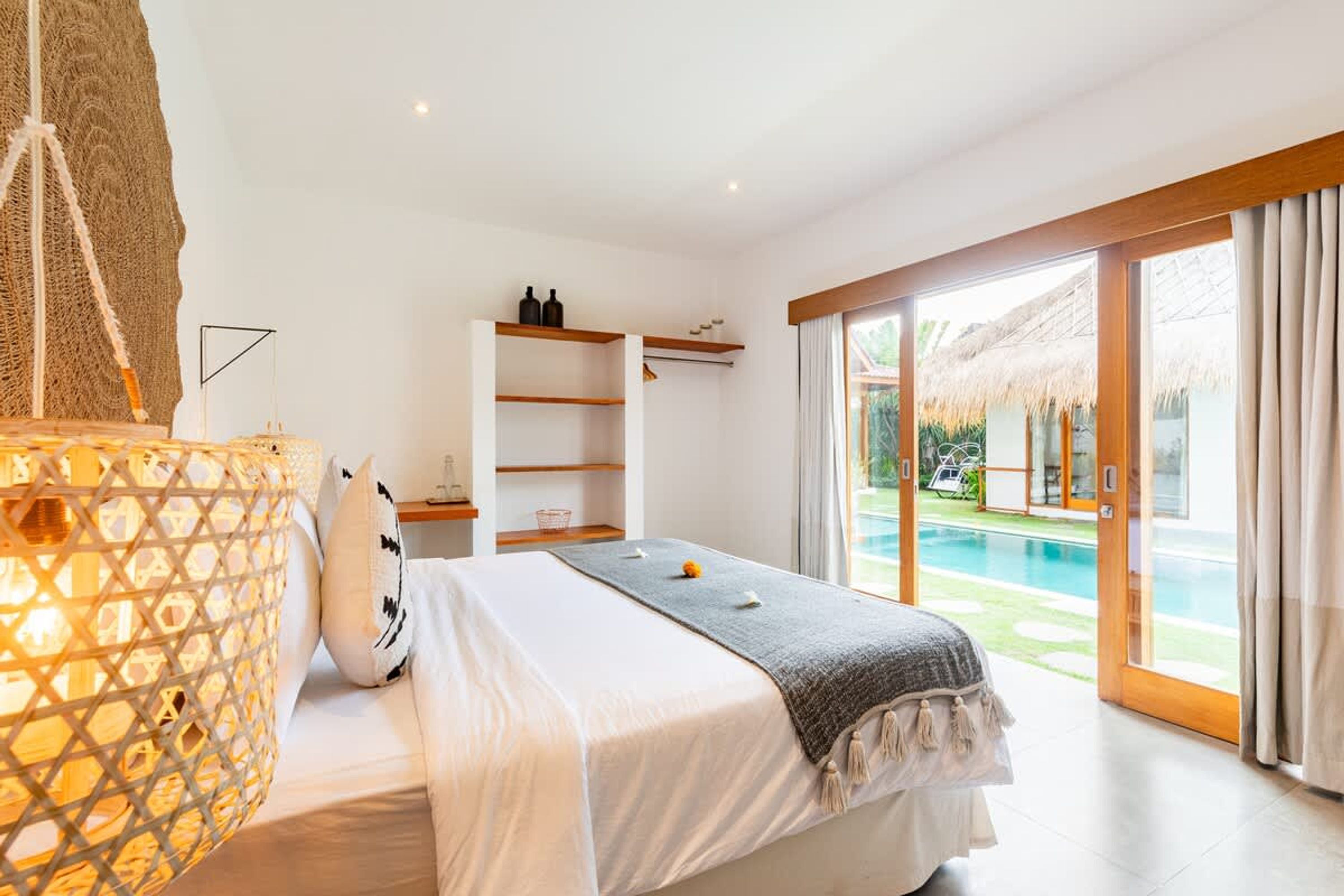 A bedroom with a bed, a nightstand, a lamp, a window with a view of a pool, and a sliding door leading to an outdoor area.