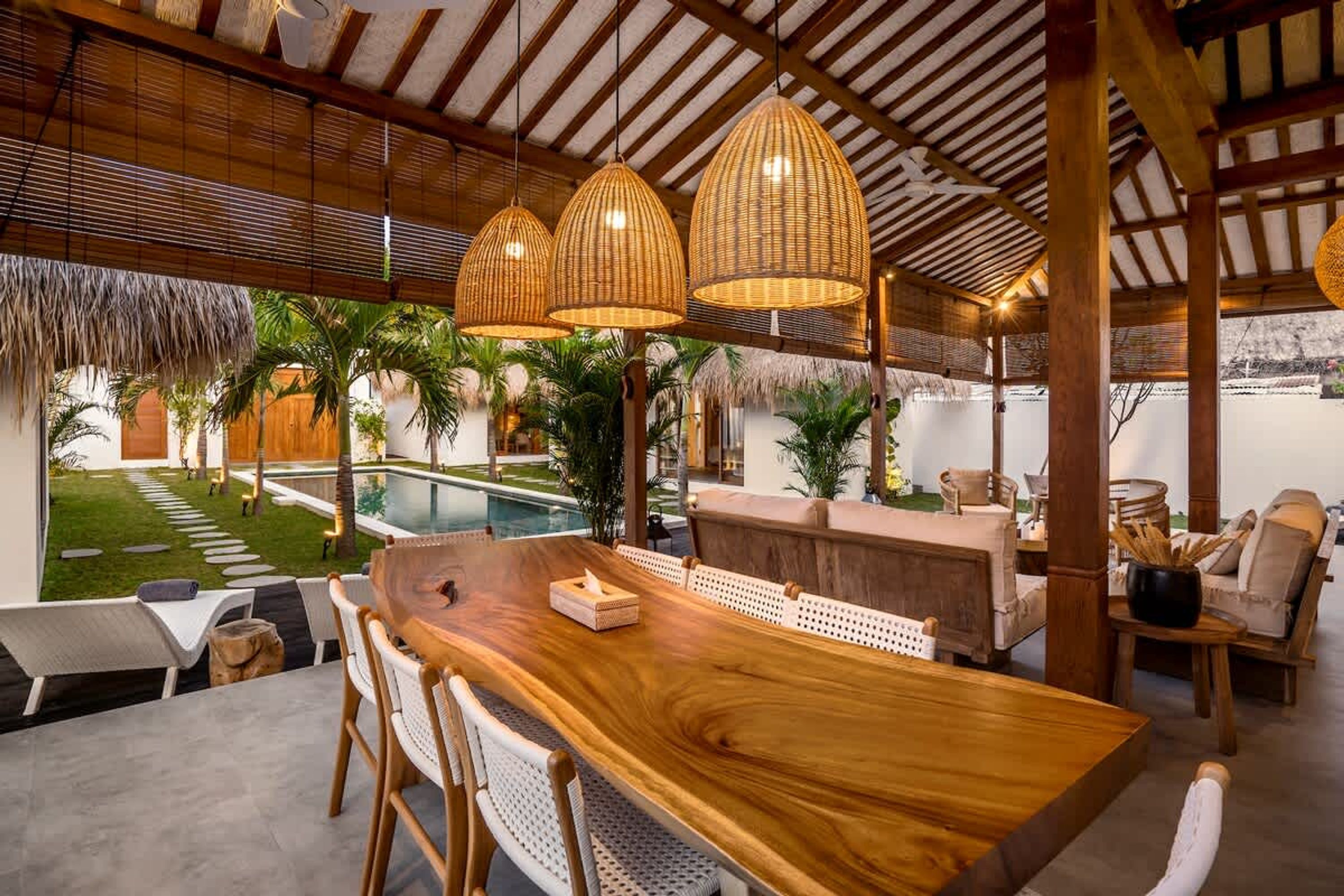 A wooden dining table with chairs, a pool, and a thatched roof structure.