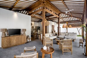 Een ruime woonkamer met een hoge nok, houten balken, een televisie, rieten meubels en een bank.