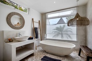 Une salle de bain moderne avec une baignoire sur pieds, un miroir rond, un meuble-lavabo blanc et une grande fenêtre donnant sur des palmiers.