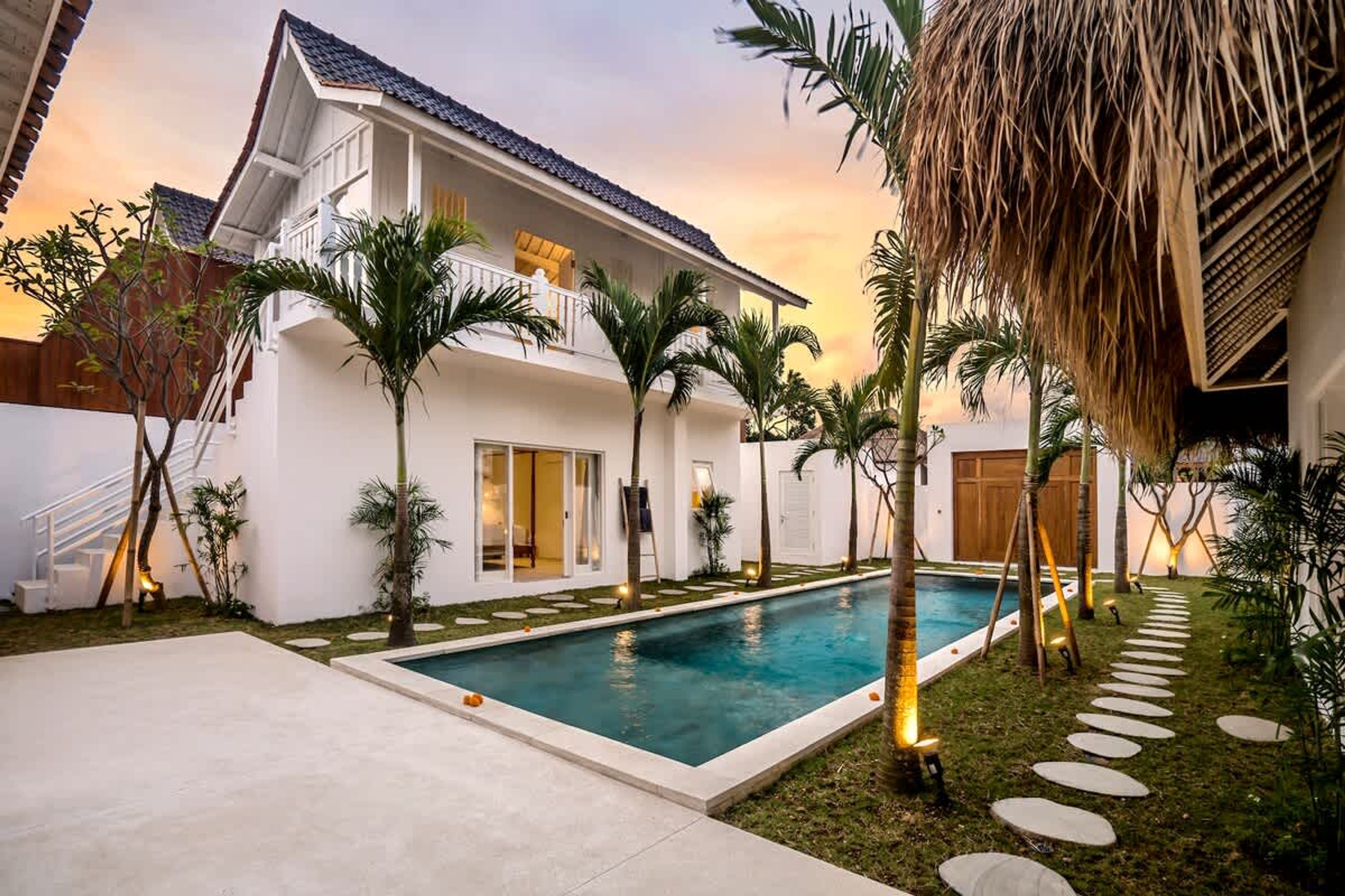 A two-story house with a swimming pool, palm trees, and a thatched roof structure.