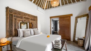 Un dormitorio amplio con una cama grande, cabecera de madera, una mesita de noche con lámpara y vistas al baño a través de una puerta.