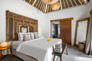 A spacious bedroom with a large bed, a wooden headboard, a bedside table with a lamp, and a view of the bathroom through a door. - Heaven 5 BR Pool Villa Seminyak Near the Beach (Dalung)