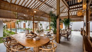 Un espacio al aire libre techado con una mesa redonda de madera y sillas de mimbre, rodeado de plantas en macetas y vistas a una piscina.