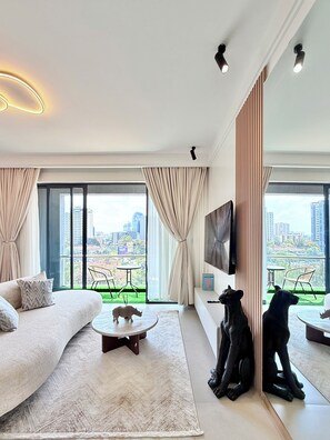 Una sala de estar moderna con sofá, mesa de centro, televisor y vista a un paisaje urbano a través de grandes ventanales.
