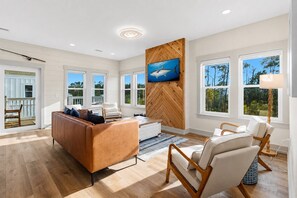 Un salón amplio con un sofá grande, una mesa de centro y dos sillones. Un televisor está montado en la pared y hay ventanas grandes que ofrecen vistas al exterior.