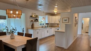 A modern kitchen with white cabinets, a wooden dining table, and a hanging light fixture. - Coastal Cottage Escape with Sunset Views, Near Beach, Bay & Wildlife Refuge (Chincoteague)