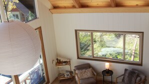 Ein gemütliches Zimmer mit Holzbalkendecke, großen Fenstern, einer Couch, zwei Sesseln, einem kleinen Tisch und einer Lampe.