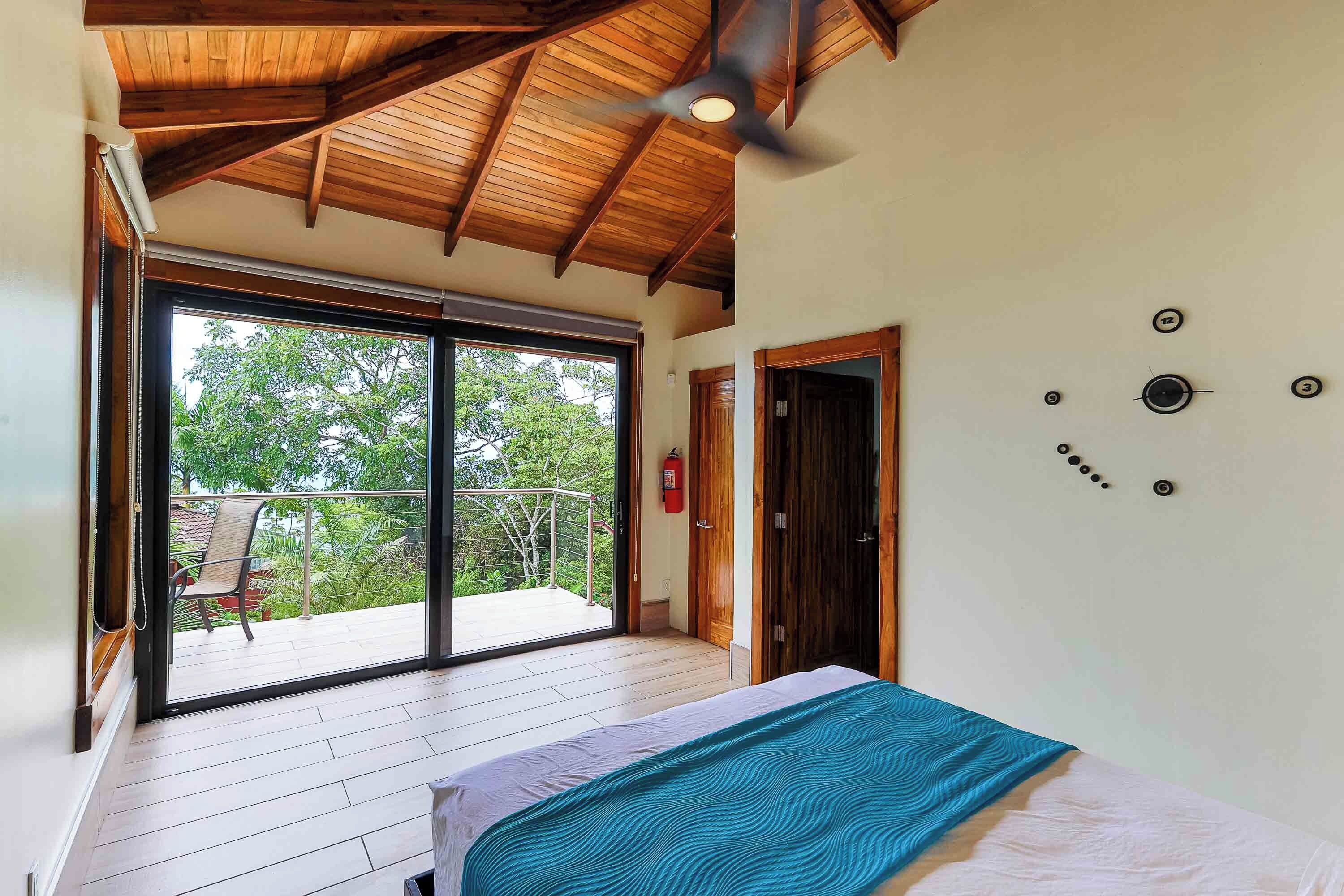 A bedroom with a bed, a ceiling fan, a large sliding glass door leading to a balcony, and a fire extinguisher on the wall.