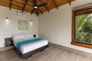 Un dormitorio con cama, ventilador de techo, ventana con vistas a vegetación y aire acondicionado de pared.