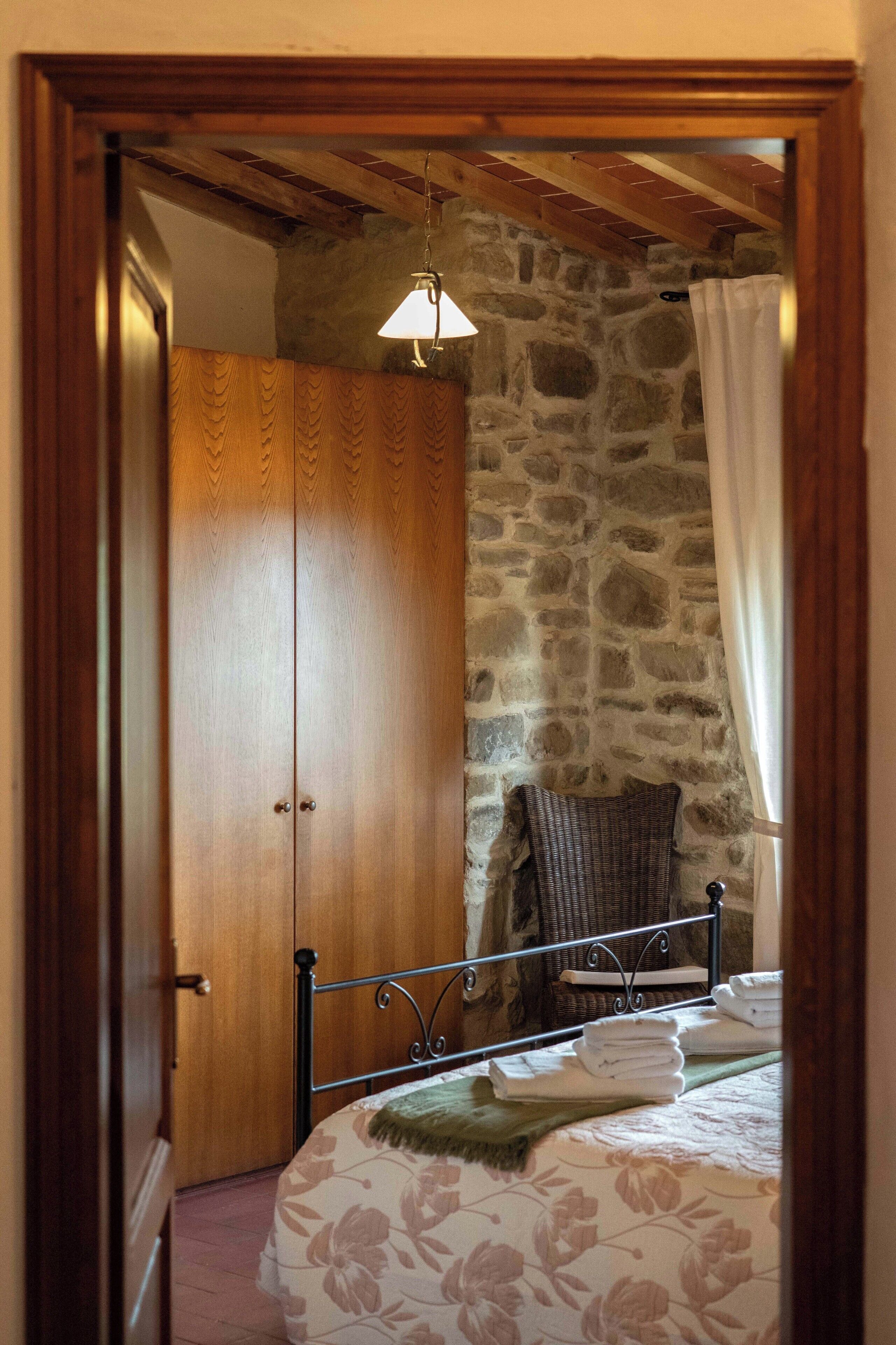 Une chambre avec une armoire en bois, un mur en pierre, un lit avec une literie à motifs floraux, une chaise en osier et une lampe suspendue.