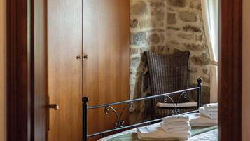 Une chambre avec une armoire en bois, un mur en pierre, un lit avec une literie à motifs floraux, une chaise en osier et une lampe suspendue.