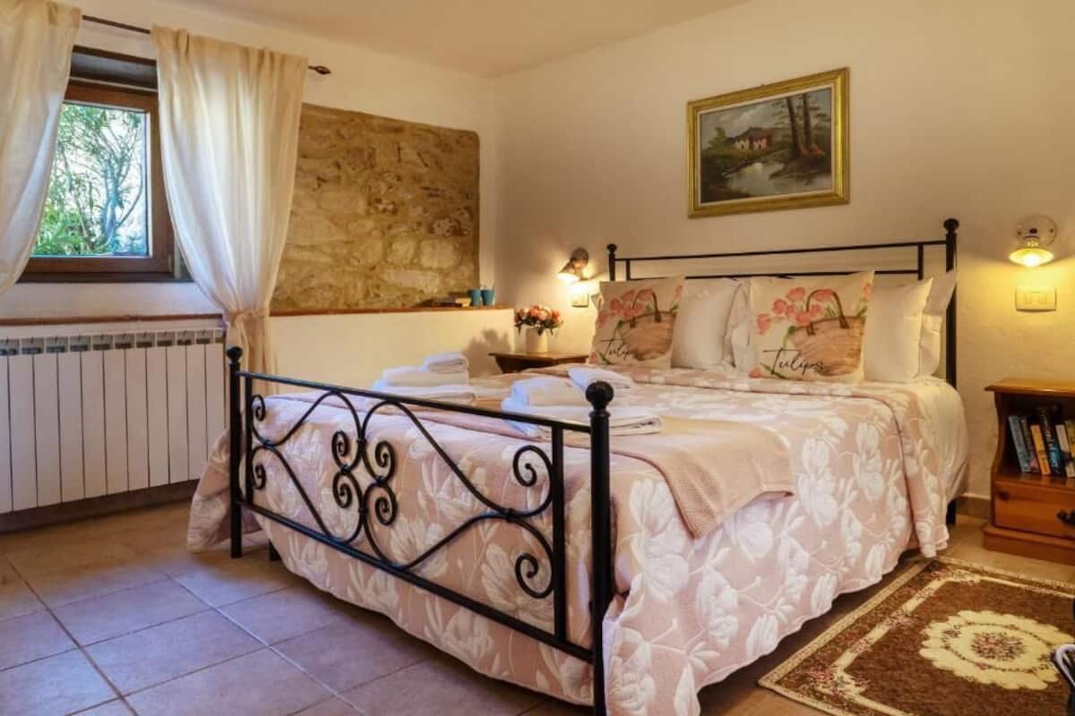 A bedroom with a bed, a stone wall, a painting, a nightstand, and a window with curtains.