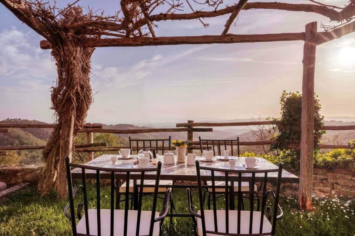 An outdoor dining area with a table set for four, surrounded by chairs, under a wooden pergola with a view of the landscape.