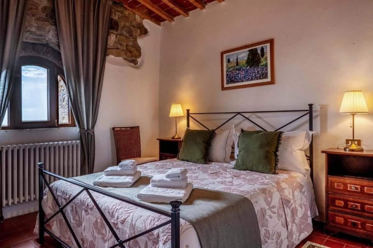 A bedroom with a bed, two bedside lamps, a radiator, a window with curtains, a picture on the wall, and a wooden chest of drawers.