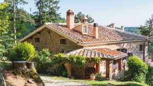 A stone house with a tiled roof, surrounded by greenery and a gravel path.