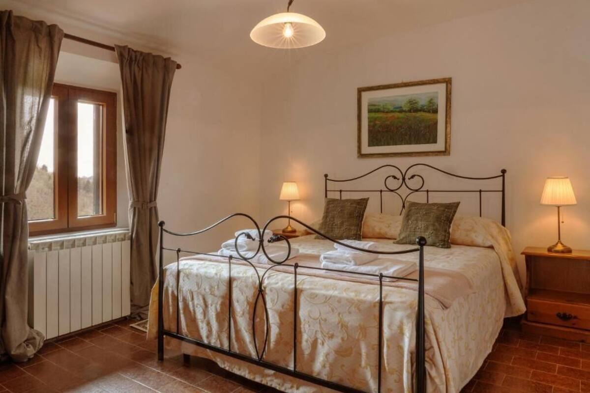 A bedroom with a bed, two bedside lamps, a window with curtains, a radiator, and a painting on the wall.
